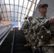 Multiple soldiers in U.S. Army camouflage (OCP/MultiCam) uniforms are riding an escalator in what appears to be a large public transit station or building with a glass-paneled ceiling. The soldiers are in full combat gear including body armor, tactical pouches, and appear to be carrying weapons. A civilian in casual clothing is visible on a parallel escalator to the left.