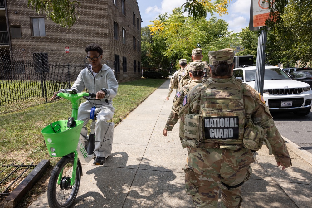 A group of National Guard soldiers in camouflage uniforms patrol a residential sidewalk in an urban neighborhood. A young civilian on a bright green Lime rental bike passes by them on the sidewalk. The scene appears to take place in a mid-rise apartment complex area on a sunny day.
