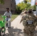 A group of National Guard soldiers in camouflage uniforms patrol a residential sidewalk in an urban neighborhood. A young civilian on a bright green Lime rental bike passes by them on the sidewalk. The scene appears to take place in a mid-rise apartment complex area on a sunny day.