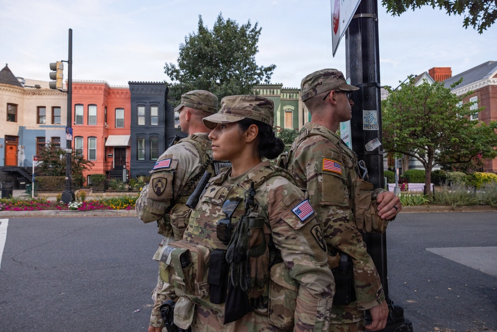 Three U.S. military personnel in full combat gear and OCP (Operational Camouflage Pattern) uniforms are standing at a street corner, appearing to be on patrol or security duty in an urban area. All three wear American flag patches on their sleeves and are equipped with tactical vests and sidearms. The setting appears to be a residential neighborhood with colorful rowhouses, consistent with Washington, D.C.