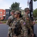 Three U.S. military personnel in full combat gear and OCP (Operational Camouflage Pattern) uniforms are standing at a street corner, appearing to be on patrol or security duty in an urban area. All three wear American flag patches on their sleeves and are equipped with tactical vests and sidearms. The setting appears to be a residential neighborhood with colorful rowhouses, consistent with Washington, D.C.