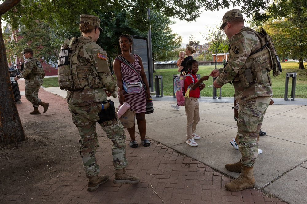 National Guard soldiers are interacting with civilians, including women and children, in an outdoor urban park or plaza setting. The soldiers appear to be distributing or reviewing documents/materials with the civilians. The setting appears calm and non-confrontational, suggesting a community outreach or assistance operation.