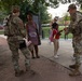 National Guard soldiers are interacting with civilians, including women and children, in an outdoor urban park or plaza setting. The soldiers appear to be distributing or reviewing documents/materials with the civilians. The setting appears calm and non-confrontational, suggesting a community outreach or assistance operation.