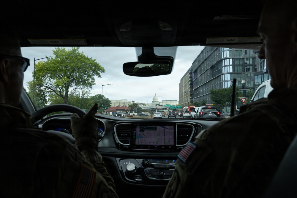 The image is taken from inside a vehicle, showing two individuals in military camouflage uniforms seated in the front seats. Through the windshield, the United States Capitol building dome is visible in the distance, indicating the vehicle is traveling through Washington, D.C. The dashboard displays an active navigation map on the infotainment screen.