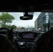 The image is taken from inside a vehicle, showing two individuals in military camouflage uniforms seated in the front seats. Through the windshield, the United States Capitol building dome is visible in the distance, indicating the vehicle is traveling through Washington, D.C. The dashboard displays an active navigation map on the infotainment screen.
