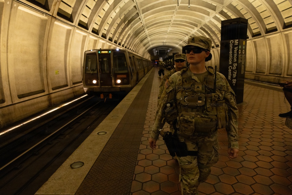 Two U.S. Army soldiers in full combat gear and camouflage uniforms are patrolling the platform of the Mt. Vernon Sq/7th St–Convention Center station on the Washington D.C. Metro system. A Metro train is visible approaching or stopped at the platform. The distinctive coffered vault architecture of the Washington Metro is clearly visible.