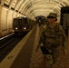 Two U.S. Army soldiers in full combat gear and camouflage uniforms are patrolling the platform of the Mt. Vernon Sq/7th St–Convention Center station on the Washington D.C. Metro system. A Metro train is visible approaching or stopped at the platform. The distinctive coffered vault architecture of the Washington Metro is clearly visible.