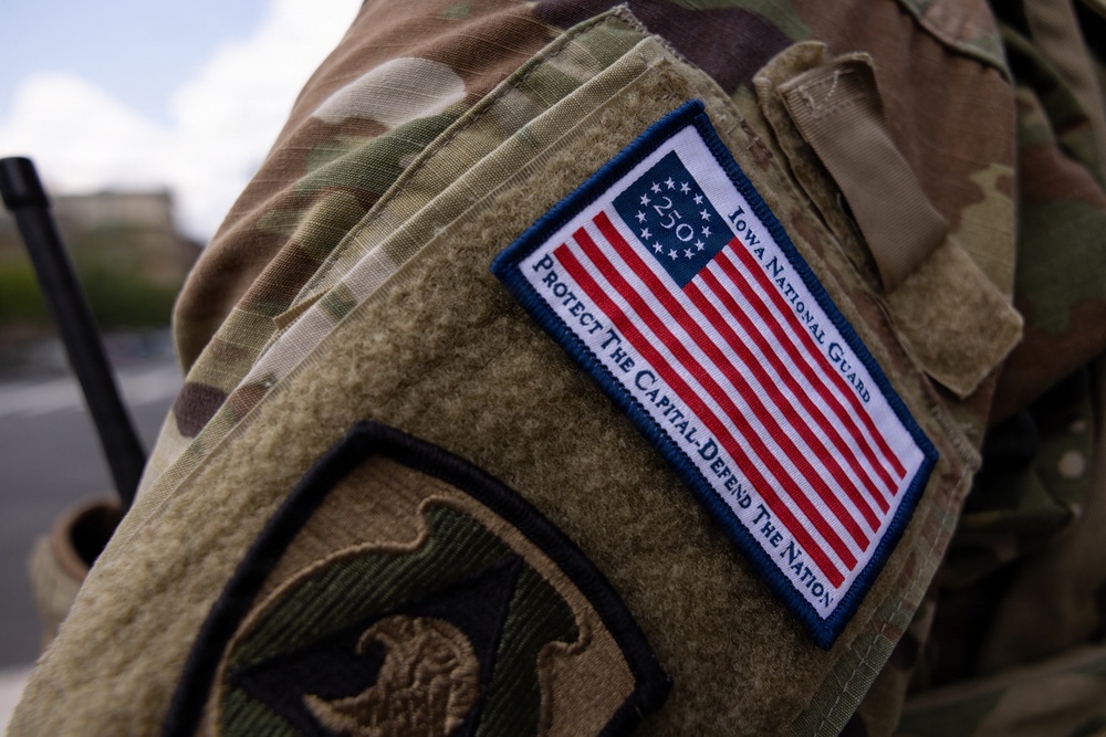 Close-up photograph of a military uniform sleeve showing a commemorative patch on OCP (Operational Camouflage Pattern) multicam fabric. The patch displays the Betsy Ross-style American flag with '250' at the center, identifying the wearer as Iowa National Guard with the motto 'Protect The Capital - Defend The Nation'. A secondary unit patch is partially visible on the lower sleeve.