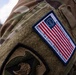 Close-up photograph of a military uniform sleeve showing a commemorative patch on OCP (Operational Camouflage Pattern) multicam fabric. The patch displays the Betsy Ross-style American flag with '250' at the center, identifying the wearer as Iowa National Guard with the motto 'Protect The Capital - Defend The Nation'. A secondary unit patch is partially visible on the lower sleeve.