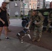 Two military personnel in camouflage uniforms interact with a civilian and his dog on a brick sidewalk in an urban residential neighborhood. One soldier is bending down to offer something to a brown and white pit bull-type dog while the owner watches. A military Humvee is parked in the background alongside a third soldier.