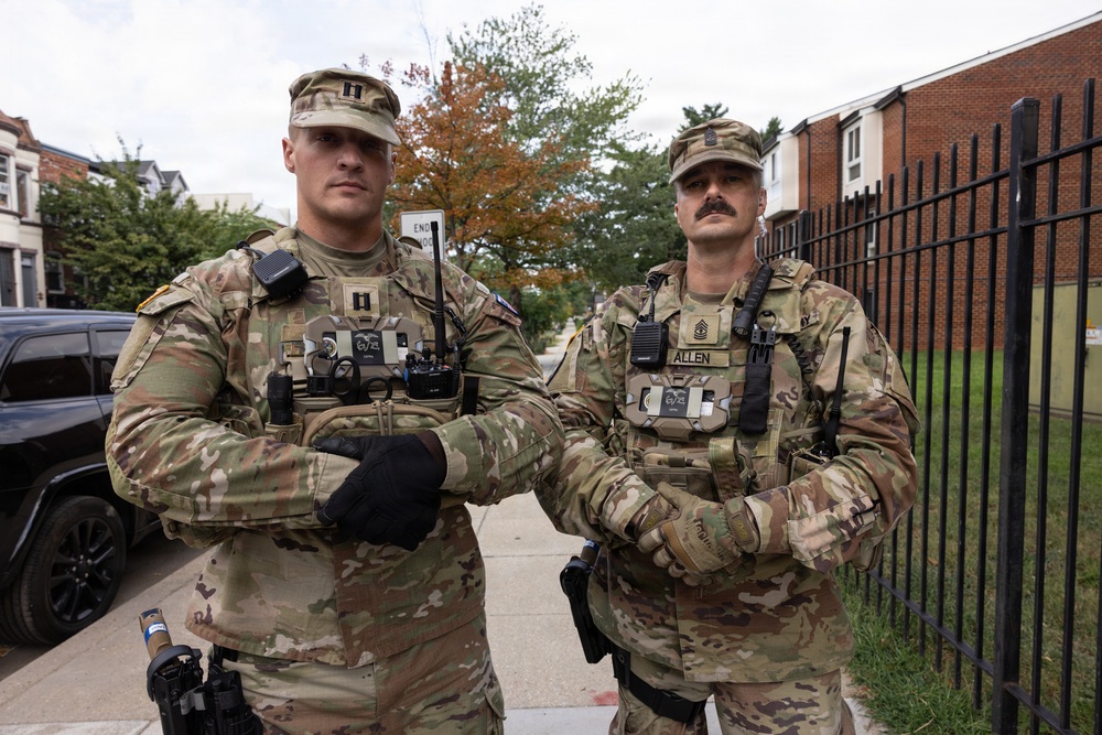 Two U.S. Army soldiers in OCP (Operational Camouflage Pattern) uniforms stand side by side on a sidewalk with arms crossed in an urban setting. The soldier on the right has a visible name tape reading 'ALLEN' and both wear rank insignia and tactical gear including radio communications equipment. They appear to be on duty in what looks like a residential or mixed-use urban area.