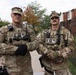 Two U.S. Army soldiers in OCP (Operational Camouflage Pattern) uniforms stand side by side on a sidewalk with arms crossed in an urban setting. The soldier on the right has a visible name tape reading 'ALLEN' and both wear rank insignia and tactical gear including radio communications equipment. They appear to be on duty in what looks like a residential or mixed-use urban area.