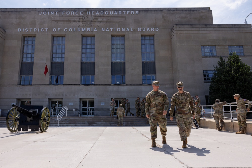 The image shows military personnel in camouflage uniforms outside the Joint Force Headquarters of the District of Columbia National Guard building. Two soldiers walk prominently in the foreground while others are visible near the building entrance. A historic artillery cannon is displayed on the left side of the plaza.