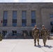 The image shows military personnel in camouflage uniforms outside the Joint Force Headquarters of the District of Columbia National Guard building. Two soldiers walk prominently in the foreground while others are visible near the building entrance. A historic artillery cannon is displayed on the left side of the plaza.