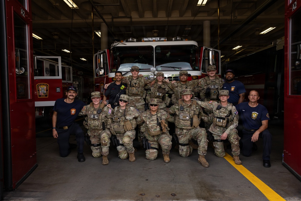 A group of approximately 13 individuals pose for a group photo in front of a red fire truck inside a fire station bay. The group consists of military personnel in camouflage (OCP/MultiCam) uniforms with tactical gear, alongside several civilian firefighters in navy blue uniforms. The setting appears to be a formal visit or joint training event between military and fire department personnel.