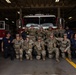 A group of approximately 13 individuals pose for a group photo in front of a red fire truck inside a fire station bay. The group consists of military personnel in camouflage (OCP/MultiCam) uniforms with tactical gear, alongside several civilian firefighters in navy blue uniforms. The setting appears to be a formal visit or joint training event between military and fire department personnel.