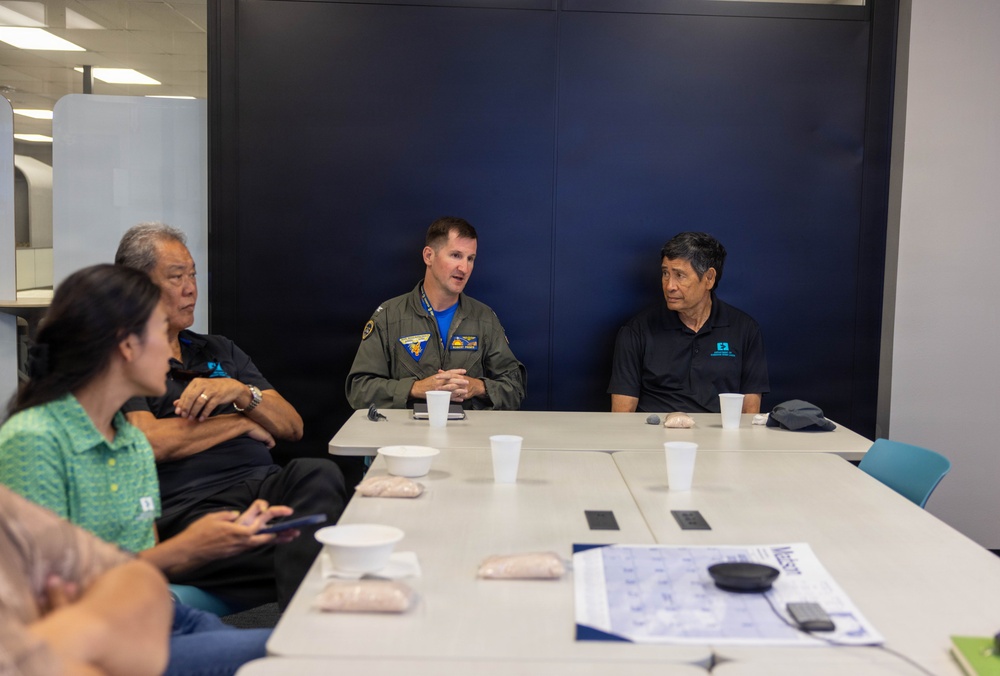 A meeting or briefing is taking place at a conference table between a military officer in a flight suit and several civilians in matching dark polo shirts. The military officer, wearing patches on his olive-green flight suit, appears to be speaking or presenting to the group. The setting appears to be a modern office or conference facility.