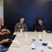 A meeting or briefing is taking place at a conference table between a military officer in a flight suit and several civilians in matching dark polo shirts. The military officer, wearing patches on his olive-green flight suit, appears to be speaking or presenting to the group. The setting appears to be a modern office or conference facility.