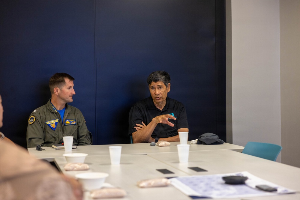 Two men are seated at a conference table in what appears to be a meeting room with a dark navy blue wall. One man is wearing a military flight suit with patches and insignia, while the other man in a black polo shirt appears to be speaking and gesturing with his hand. The table has cups, papers, phones, and snack items on it, suggesting an ongoing meeting or briefing.