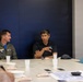 Two men are seated at a conference table in what appears to be a meeting room with a dark navy blue wall. One man is wearing a military flight suit with patches and insignia, while the other man in a black polo shirt appears to be speaking and gesturing with his hand. The table has cups, papers, phones, and snack items on it, suggesting an ongoing meeting or briefing.