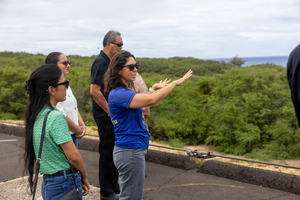 A group of four people stand on what appears to be an elevated outdoor platform or rooftop overlooking a lush green landscape with the ocean visible in the background. A woman in a blue shirt appears to be leading a tour or briefing, gesturing outward to point something out to the group. The setting suggests a coastal location, possibly Hawaii based on the vegetation and ocean backdrop.