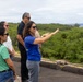 A group of four people stand on what appears to be an elevated outdoor platform or rooftop overlooking a lush green landscape with the ocean visible in the background. A woman in a blue shirt appears to be leading a tour or briefing, gesturing outward to point something out to the group. The setting suggests a coastal location, possibly Hawaii based on the vegetation and ocean backdrop.