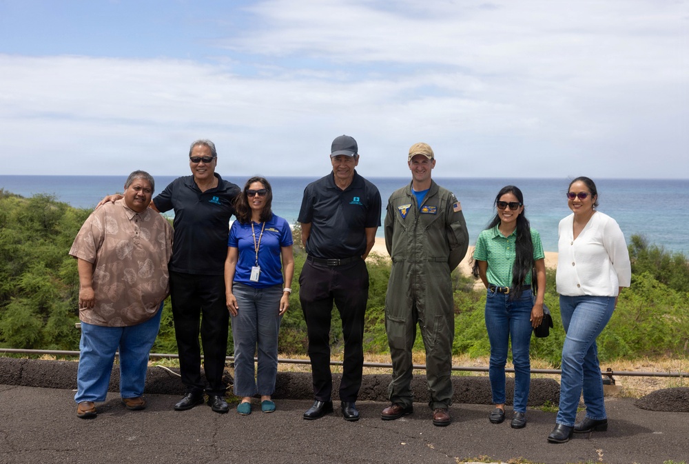 A group of seven people pose together outdoors near a coastal overlook with a beach and ocean visible in the background. The group includes individuals in civilian attire, one person in a blue polo with a lanyard/badge, and one person in a military flight suit with patches. The setting appears to be a scenic overlook in a tropical or subtropical location, likely Hawaii based on the vegetation and coastline.
