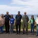 A group of seven people pose together outdoors near a coastal overlook with a beach and ocean visible in the background. The group includes individuals in civilian attire, one person in a blue polo with a lanyard/badge, and one person in a military flight suit with patches. The setting appears to be a scenic overlook in a tropical or subtropical location, likely Hawaii based on the vegetation and coastline.