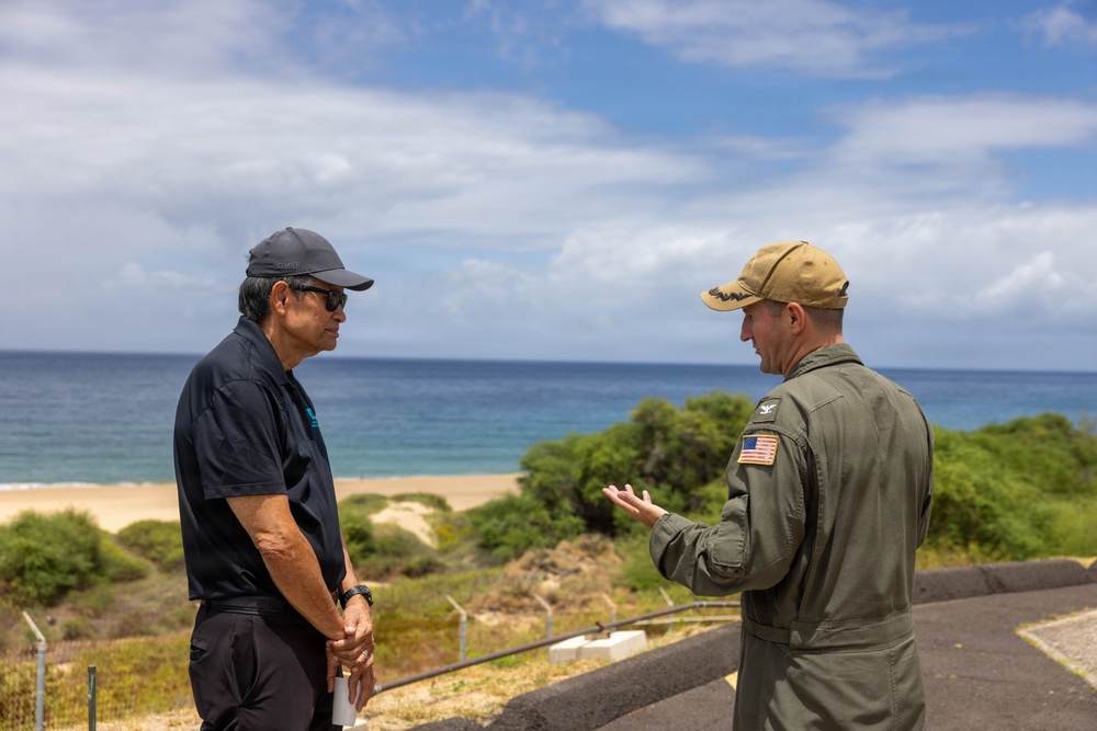 Two men are engaged in conversation outdoors at what appears to be a coastal military or government installation. One man is wearing a U.S. military flight suit with an American flag patch and a tan cap with what appears to be a military insignia, while the other is dressed in civilian attire with a dark polo shirt and baseball cap. The backdrop features a sandy beach, ocean, and partially cloudy sky.