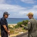 Two men are engaged in conversation outdoors at what appears to be a coastal military or government installation. One man is wearing a U.S. military flight suit with an American flag patch and a tan cap with what appears to be a military insignia, while the other is dressed in civilian attire with a dark polo shirt and baseball cap. The backdrop features a sandy beach, ocean, and partially cloudy sky.