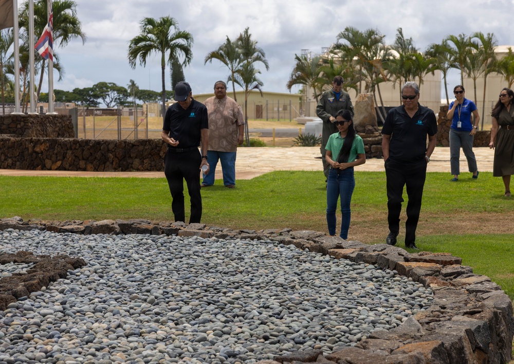 A group of people are walking around an outdoor site featuring a large stone and gravel structure, likely a Hawaiian heiau (traditional temple platform) or cultural monument. The setting appears to be on a military installation in Hawaii, evidenced by the Hawaiian state flag visible on a flagpole and institutional buildings in the background. Several individuals wearing matching dark polo shirts with a logo suggest an organized official visit or tour.