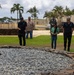 A group of people are walking around an outdoor site featuring a large stone and gravel structure, likely a Hawaiian heiau (traditional temple platform) or cultural monument. The setting appears to be on a military installation in Hawaii, evidenced by the Hawaiian state flag visible on a flagpole and institutional buildings in the background. Several individuals wearing matching dark polo shirts with a logo suggest an organized official visit or tour.