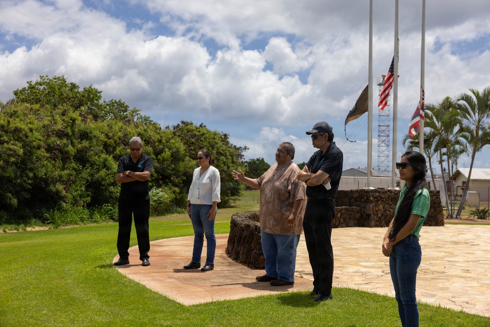 Five individuals are gathered outdoors on a stone-paved area surrounded by well-maintained green lawn, appearing to be at an official or memorial site. One person in the center appears to be speaking or presenting to the group, gesturing with their hands. Multiple flagpoles are visible in the background, with what appears to be an American flag and a Hawaiian state flag flying.
