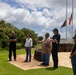 Five individuals are gathered outdoors on a stone-paved area surrounded by well-maintained green lawn, appearing to be at an official or memorial site. One person in the center appears to be speaking or presenting to the group, gesturing with their hands. Multiple flagpoles are visible in the background, with what appears to be an American flag and a Hawaiian state flag flying.