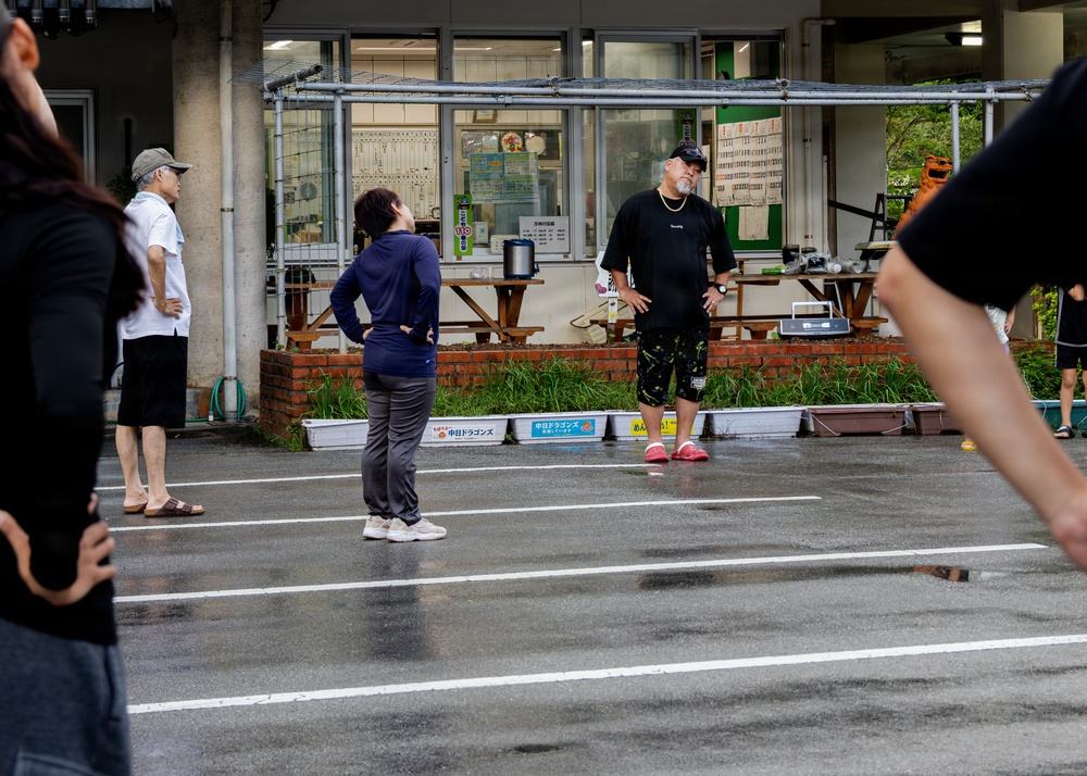 A group of people are gathered in a wet parking lot outside a building with Japanese signage, suggesting recent rainfall. A tall man in a black shirt, dark patterned shorts, and pink shoes appears to be instructing or addressing a woman in a navy blue outfit. The setting appears to be in Japan, with visible Japanese text on banners and storefront signs.
