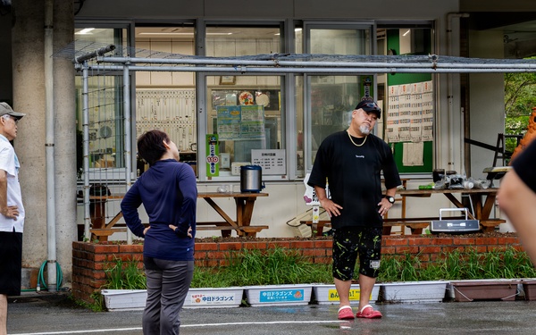 A group of people are gathered in a wet parking lot outside a building with Japanese signage, suggesting recent rainfall. A tall man in a black shirt, dark patterned shorts, and pink shoes appears to be instructing or addressing a woman in a navy blue outfit. The setting appears to be in Japan, with visible Japanese text on banners and storefront signs.