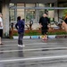 A group of people are gathered in a wet parking lot outside a building with Japanese signage, suggesting recent rainfall. A tall man in a black shirt, dark patterned shorts, and pink shoes appears to be instructing or addressing a woman in a navy blue outfit. The setting appears to be in Japan, with visible Japanese text on banners and storefront signs.