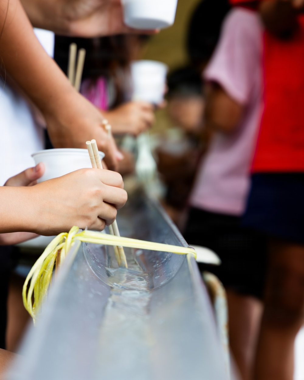 The image shows people participating in a traditional Japanese nagashi somen (flowing noodles) activity, where yellow-green somen noodles flow through a water-filled bamboo or plastic chute. Participants are using chopsticks to catch the noodles as they pass by, while holding small white cups. Multiple people of varying ages are visible along the length of the chute.