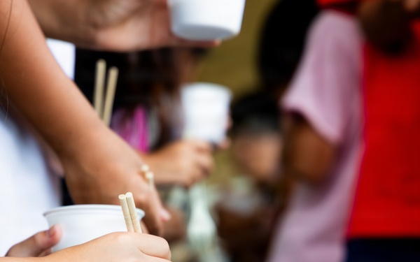 The image shows people participating in a traditional Japanese nagashi somen (flowing noodles) activity, where yellow-green somen noodles flow through a water-filled bamboo or plastic chute. Participants are using chopsticks to catch the noodles as they pass by, while holding small white cups. Multiple people of varying ages are visible along the length of the chute.