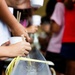 The image shows people participating in a traditional Japanese nagashi somen (flowing noodles) activity, where yellow-green somen noodles flow through a water-filled bamboo or plastic chute. Participants are using chopsticks to catch the noodles as they pass by, while holding small white cups. Multiple people of varying ages are visible along the length of the chute.
