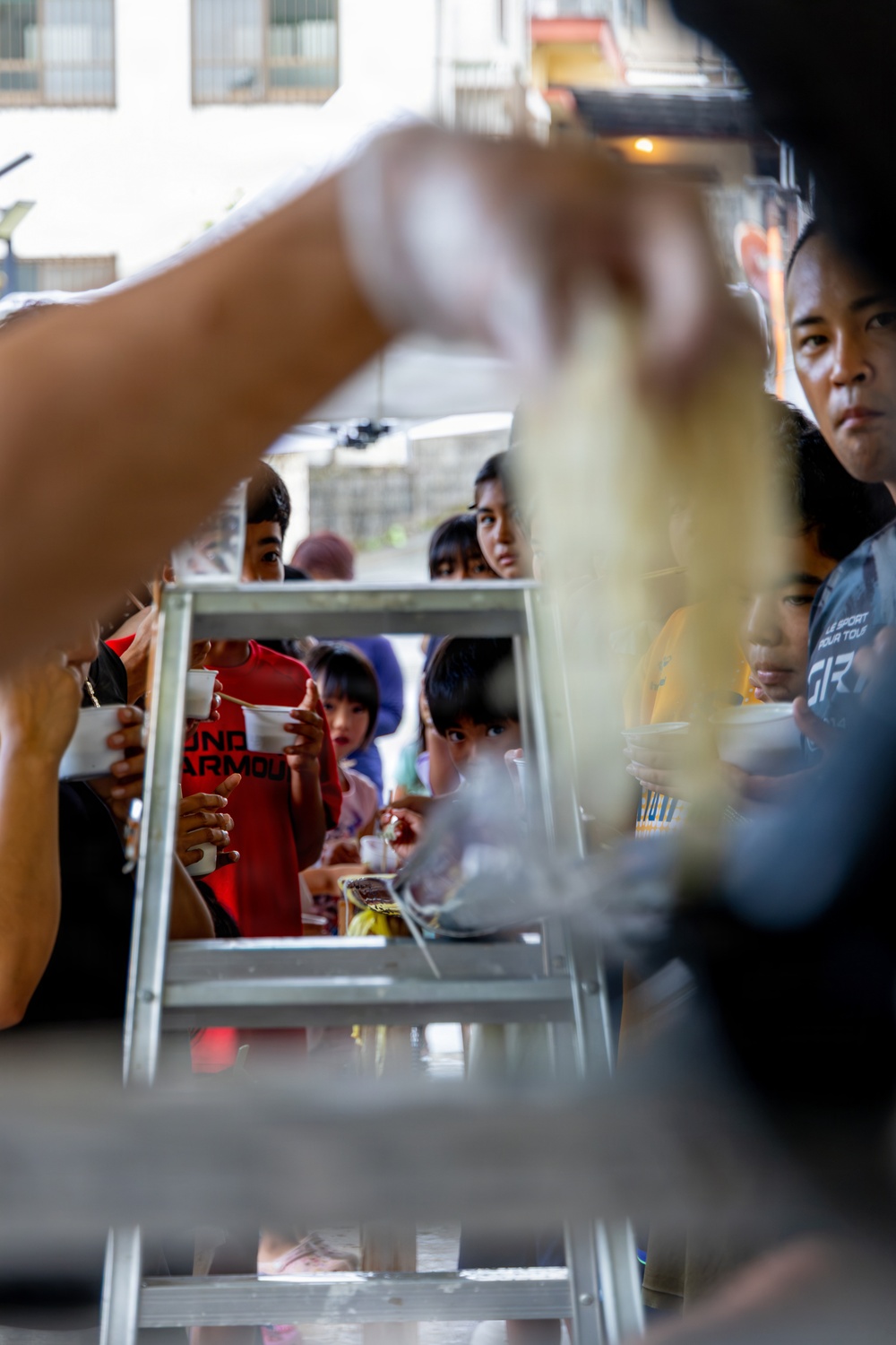 A food distribution or feeding event is taking place, with multiple children and adults gathered and holding small cups or bowls. The scene is photographed from behind a serving counter or table, with a hand in the foreground appearing to ladle or pour food. The crowd appears to be waiting to receive food, suggesting a community outreach or charity feeding program.