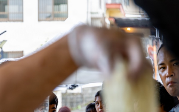 A food distribution or feeding event is taking place, with multiple children and adults gathered and holding small cups or bowls. The scene is photographed from behind a serving counter or table, with a hand in the foreground appearing to ladle or pour food. The crowd appears to be waiting to receive food, suggesting a community outreach or charity feeding program.