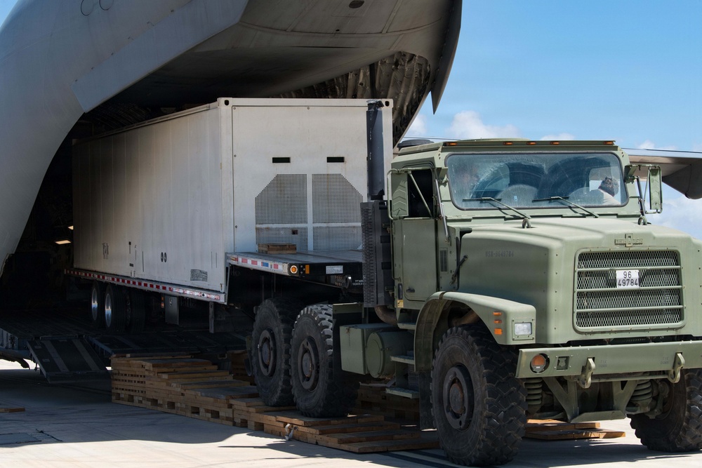 A military olive-drab heavy truck is being used to load or unload a large white trailer/container from the rear cargo ramp of a military transport aircraft. The operation is taking place on a tarmac under clear blue skies. Wooden dunnage boards are visible on the ground beneath the truck's wheels to support the ramp transition.