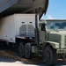 A military olive-drab heavy truck is being used to load or unload a large white trailer/container from the rear cargo ramp of a military transport aircraft. The operation is taking place on a tarmac under clear blue skies. Wooden dunnage boards are visible on the ground beneath the truck's wheels to support the ramp transition.