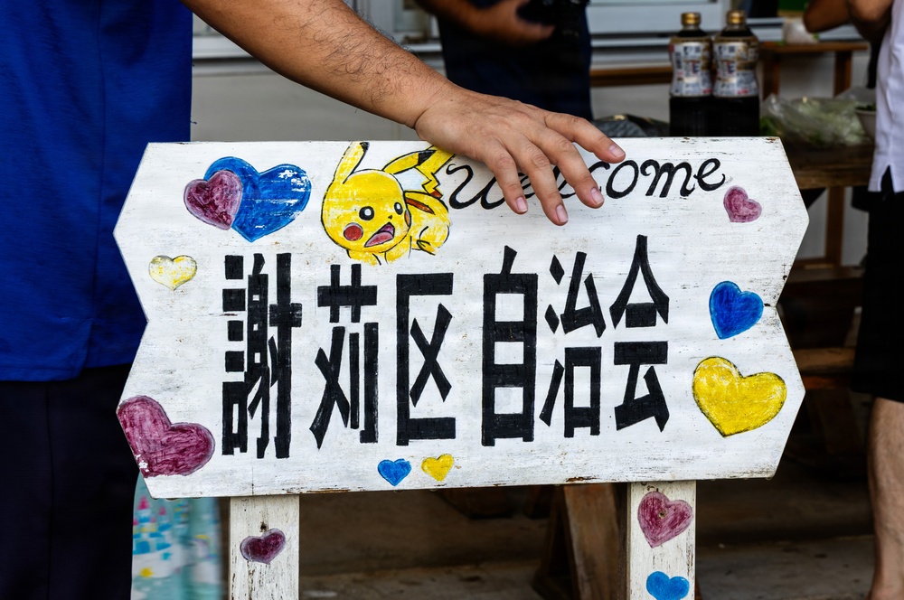 A hand-painted wooden sign featuring Japanese/Chinese characters (謝莉区自治会, meaning approximately 'Sheri District Residents' Association'), decorated with a Pikachu illustration and colorful hand-drawn hearts. The word 'welcome' is partially visible at the top. A person in a blue shirt is holding or steadying the sign.