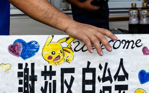 A hand-painted wooden sign featuring Japanese/Chinese characters (謝莉区自治会, meaning approximately 'Sheri District Residents' Association'), decorated with a Pikachu illustration and colorful hand-drawn hearts. The word 'welcome' is partially visible at the top. A person in a blue shirt is holding or steadying the sign.