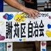 A hand-painted wooden sign featuring Japanese/Chinese characters (謝莉区自治会, meaning approximately 'Sheri District Residents' Association'), decorated with a Pikachu illustration and colorful hand-drawn hearts. The word 'welcome' is partially visible at the top. A person in a blue shirt is holding or steadying the sign.