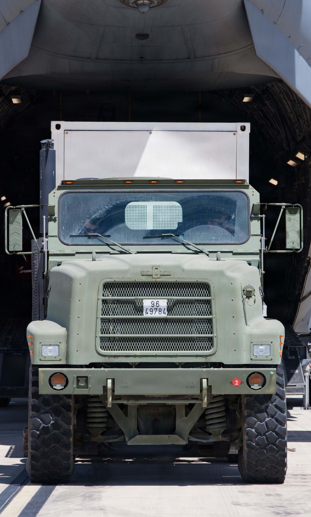 A military olive-drab truck is positioned at the open rear cargo ramp of a large military transport aircraft, appearing to be in the process of loading or unloading. The truck bears a military registration plate reading '96 49784' and displays a red number '2' marker on its front bumper. The scene is photographed from a direct front-facing perspective in bright daylight conditions.