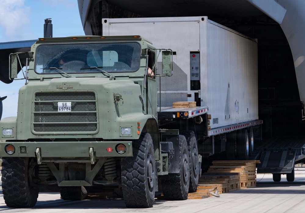 A military heavy-duty truck (olive drab/green) is pulling a flatbed trailer loaded with a large white equipment shelter or container out of the rear cargo bay of a military transport aircraft. The operation appears to be an offloading or unloading exercise on an airfield tarmac. Wooden dunnage/planks are visible on the ground, used to manage the ramp transition.