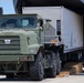 A military heavy-duty truck (olive drab/green) is pulling a flatbed trailer loaded with a large white equipment shelter or container out of the rear cargo bay of a military transport aircraft. The operation appears to be an offloading or unloading exercise on an airfield tarmac. Wooden dunnage/planks are visible on the ground, used to manage the ramp transition.
